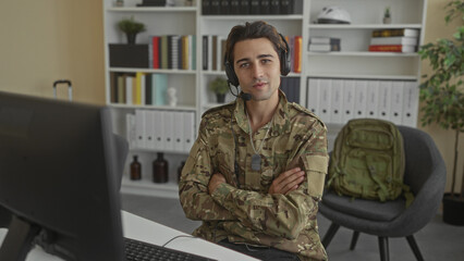 Fototapeta premium Soldier man typing with headset, hands on keyboard and dog tags visible while using a desktop computer at a desk with bookshelves and a backpack in an office building; duty.