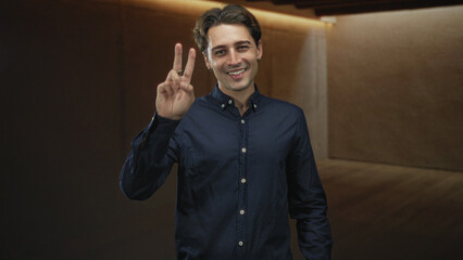 Obraz premium Man in navy shirt smiling and holding up peace sign with fingers in studio; optimism friendliness welcome connection.