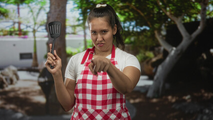 Young woman in red checkered apron holding a spatula and pointing finger at camera in forest setting near trees and picnic area  defiance cooking. © Krakenimages.com