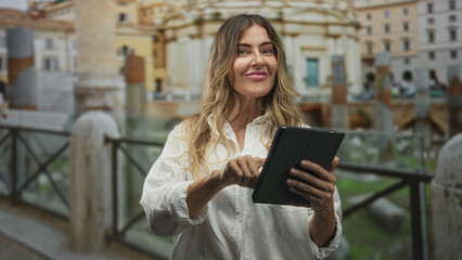 Woman taps tablet with right finger at stone railing in front of historic building and fountain, smiling and looking at screen  serenity curiosity. © Krakenimages.com