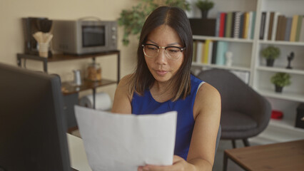 Fototapeta premium Woman reading a paper document at a desk in an office building, holding the sheet near a computer monitor and wearing glasses while seated at work; concentration productivity.