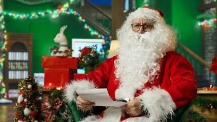 Fototapeta premium Elderly man dressed as Santa Claus opening letter received from kids asking for presents. Older person wearing Santa costume in Christmas decorated room reading letter from children, camera A
