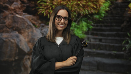 Woman judge wearing glasses and black robe holding a gavel upright with one hand, smiling before stone steps in studio  confidence justice. © Krakenimages.com
