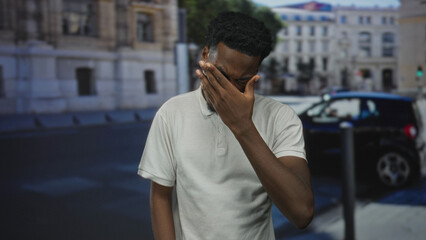 Fototapeta premium Man wipes tear from eye on street in morning light with hand touching eye and subtle grimace; sadness.
