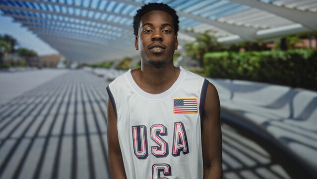 Man in usa basketball jersey and american flag patch extends open palm gesture on sunlit modern building walkway; uncertainty.