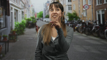 Fototapeta premium Young woman points index finger while smiling, finger to lips in shh pose, wearing gray cardigan on a cobblestone street lined with bicycles and brick buildings; playful secret.