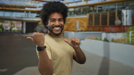 Smiling man with a beard points thumbs behind in a city street scene, wearing casual clothing,...