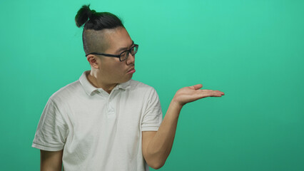 Fototapeta premium Man with glasses and topknot holding palms up gesture in green studio; confusion pondering choice dilemma.