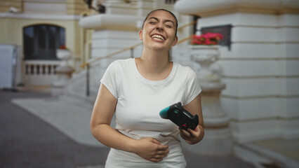 Woman holding gamepad laughing with head thrown back in front of ornate building entrance, casual white t shirt, right hand gripping controller and left hand on stomach  amusement. © Krakenimages.com