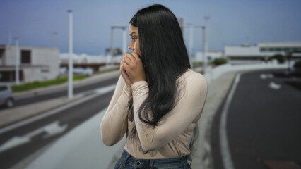 Young latin woman stands alone clasped hands near face on street under clear daylight  anxiety vulnerability uncertainty. © Krakenimages.com