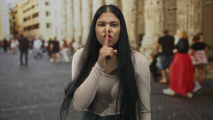 Fototapeta premium Young latin woman finger to lips for silence on cobblestone street beside stone building; tranquility.