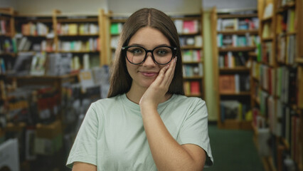 Teenage girl touches her glasses by temple amid wooden bookshelves and books in a library building  confidence. © Krakenimages.com