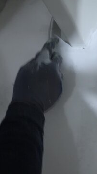 Macro close up of a worker&rsquo;s hand using a putty knife to remove imperfections from wall and metal edges. Renovation detail shows surface preparation work.