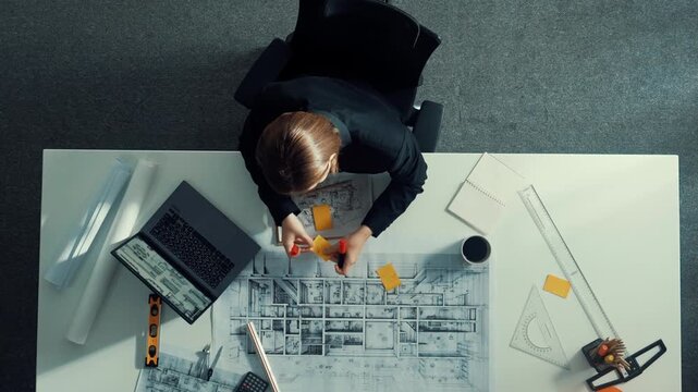 Aerial view of architect drawing while working at laptop. Top down view of caucasian engineer designing blueprint and drafting house model at table with equipment and safety helmet. Alimentation.