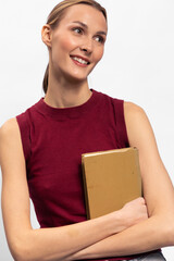 Obraz premium Smiling young woman in sleeveless red top holds a cardboard box while looking away. Studio portrait on white background with casual posture and positive mood. Concept of delivery and packaging.