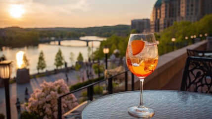 Fototapeta premium Glass of refreshing orange cocktail rests on an outdoor patio table overlooking a cityscape and river at sunset