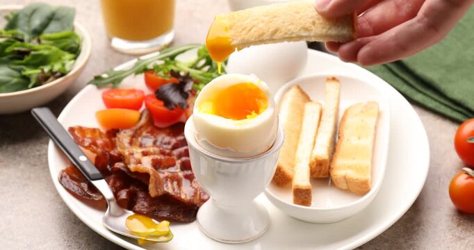 Tasty breakfast. Woman dipping toasted bread into soft boiled egg at grey table, closeup