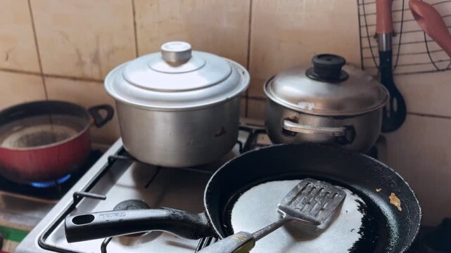 Close view of aluminum pots and nonstick frying pan on a lit gas stove, spatula resting in pan, tiled backsplash and utensils, everyday home kitchen cooking setup.
