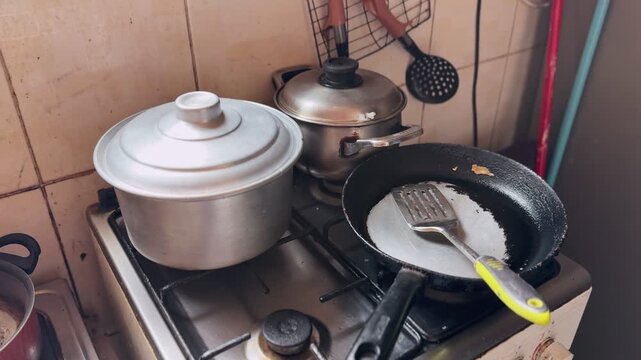 Wide view of lidded pots and an empty worn frying pan with spatula on a stainless gas stove, tiled backsplash and hanging utensils, showing everyday domestic cooking setup.