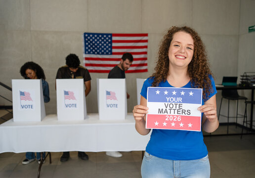 caucasian American woman smiles holding a Your Vote Matters 2026 sign at a US election polling station. In the background, a diverse group of people votes in voting booths, democracy in America