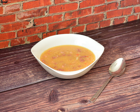 Pea soup with smoked meat in a deep plate on a wooden table, a tablespoon nearby.