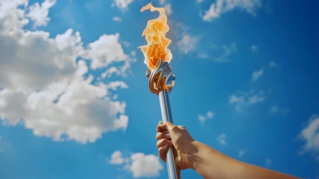 Hand holding torch against blue sky, bright clouds, upward angle, daytime light, metal pole gripped firmly, roaring orange flame, ceremonial atmosphere conveying unity, celebration,