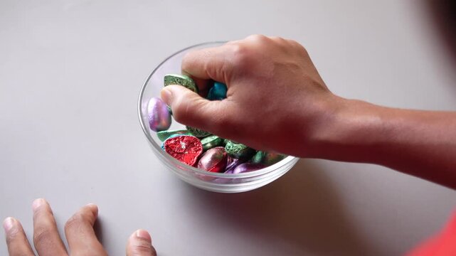 Child's hand reaching into bowl of colorful heart-shaped chocolates, grabbing handful of sweet candies from above shot, capturing moment of indulgence