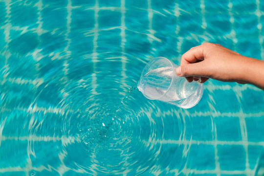 Pouring liquid pool chemicals from a transparent measuring cup into clear blue swimming pool water during routine maintenance and water treatment process
