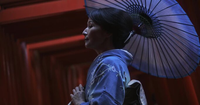 Beautiful Japanese woman in kimono with umbrella standing in shadows of ancient torii gates - slow motion