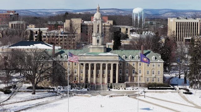 Aerial view of Penn State University&rsquo;s Old Main building covered in fresh snow, with American flags waving in front of historic campus landmark on a bright winter day. Wide shot.
