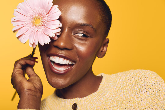 Happy smiling African woman with dark skin holds pink flower near face against yellow background. Concept of beauty, joy, skincare, natural makeup, and positive emotions in studio.