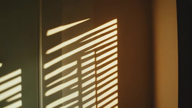 Sunlight streams through window blinds, casting a repeating pattern of bright stripes and dark shadows across an interior wall, forming a geometric design during the golden hour