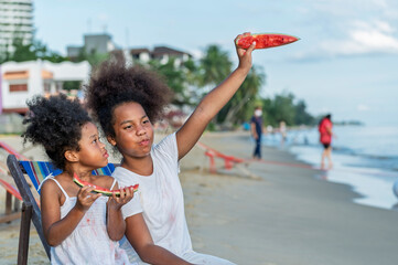 Two smiling African American girls with curly hair bite watermelon and share a happy moment of pure beauty and fun in Beach outdoors background