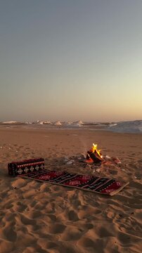 Vertical shot of desert camping scene with red arabian carpet on the sand and bonfire at sunset