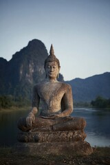 Obraz premium Ancient Stone Buddha Statue Meditating by a Peaceful River with Mountain Backdrop for Spiritual Tourism, Wellness Brochures, and Tranquil Morning Background with Copy Space