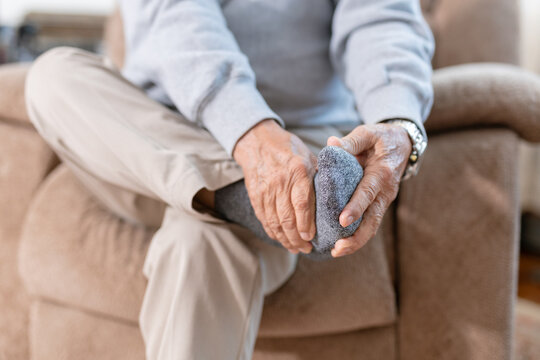 Senior Asian man suffering from foot pain at home, holding his heel while sitting on sofa. Elderly male with plantar fasciitis, foot cramp, healthcare and medical concept.