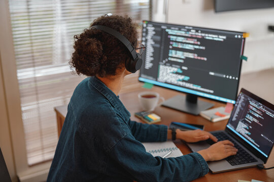 Remote software developer wearing headphones, writing and testing code on dual screens at a cozy home office desk with coffee and notes nearby

