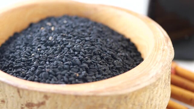 Close up view of nigella seeds in wooden bowl with bottle of oil and cinnamon sticks in background, black cumin seeds used as spice and traditional medicine 