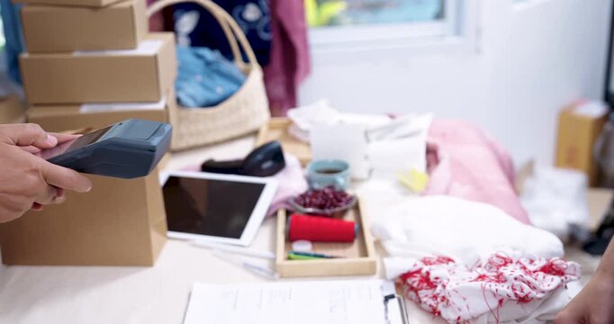 Retired asian woman artisan slides credit card near EDC machine, checks invoice beside laptop, boxes ready for shipping, craft entrepreneur processes order fulfillment, shibori textile delivery.