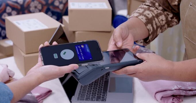 Retired asian woman entrepreneur angles EDC machine toward customer smartphone, confirmation check appears, boxes stacked by laptop, cashless transaction supports handmade shibori textile shipment