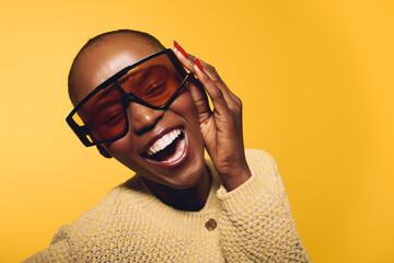 Smiling dark skinned African woman wearing oversized sunglasses and beige sweater poses joyfully against yellow background. Concept of happiness, fashion, and confident black female model.