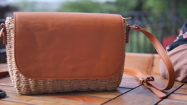 A static close-up shot of a chic women's handbag made of woven straw and brown leather, sitting on a rustic wooden table with a gentle bokeh background, capturing its intricate details and texture 