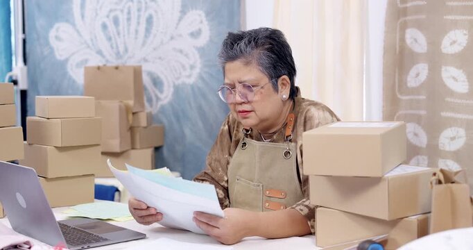 Older asian woman entrepreneur studies invoice pages, checks laptop order record, compares receipt lines, sorts paperwork bundle, plans packing count, prepares shipping schedule inside home studio.
