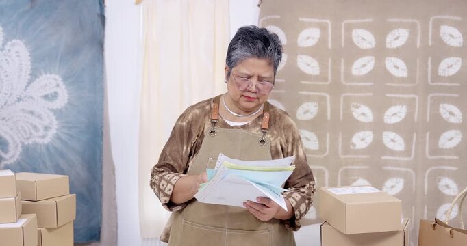 Retired artisan businesswoman reads invoice packet, checks receipt details, updates order list on laptop, holds paperwork bundle, plans parcel shipping route, prepares packing inside home studio.