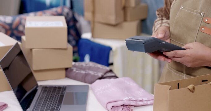 Asian female businesswoman accepts smartwatch tap to pay on POS terminal, retired woman small business prepares sustainable slow fashion order, checks invoice, seals handmade shibori fabric parcel.