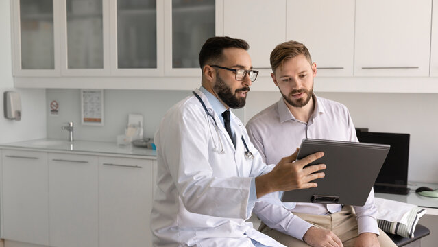 Discussing health report. Male doctor sit on couch with young patient visiting clinic show medical document on clipboard explain therapy schedule present diet plan review checkup results. Copy space