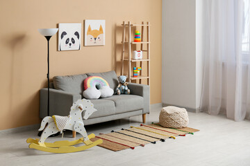 Interior of children's room with sofa, lamp, many toys in shelving unit and rocking horse near beige wall