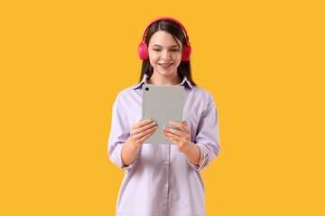 Beautiful young woman in headphones with modern tablet computer on yellow background
