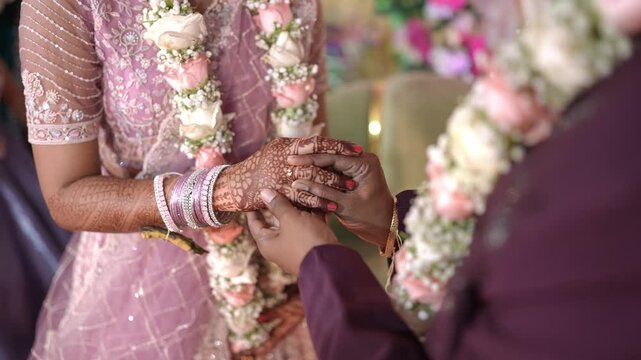 Cinematic close-up of a groom placing a gold engagement ring on the bride's henna-adorned hand during a traditional Indian wedding ceremony with pastel floral garlands.