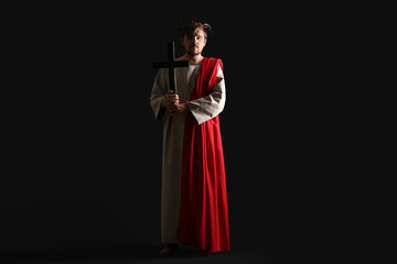 Fototapeta premium Man in Jesus robe and crown of thorns holding wooden cross with light on black background